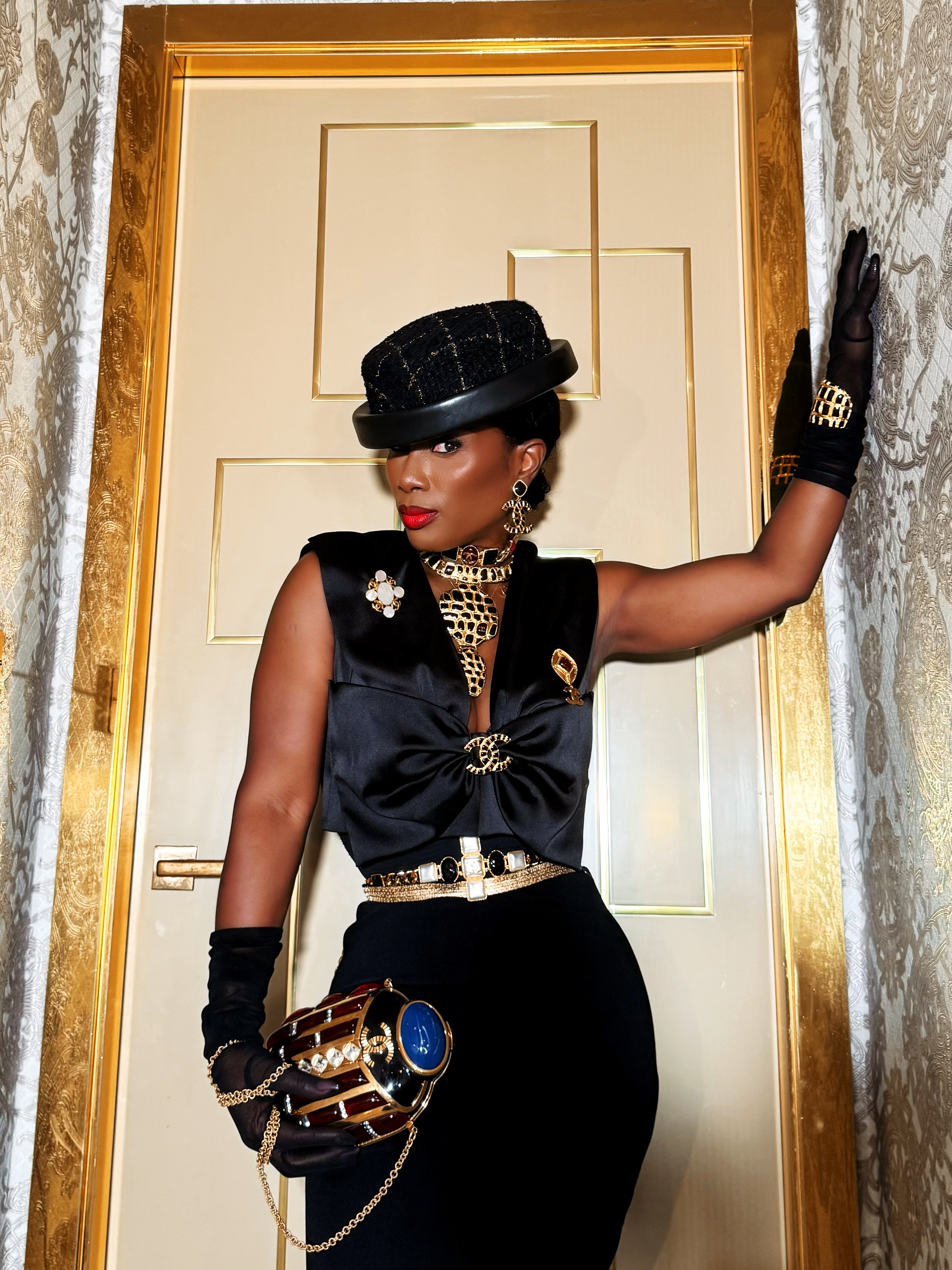 Dede Chancelor in black Chanel — hat, gloves, gold jewellery, leaning against a gilded door.