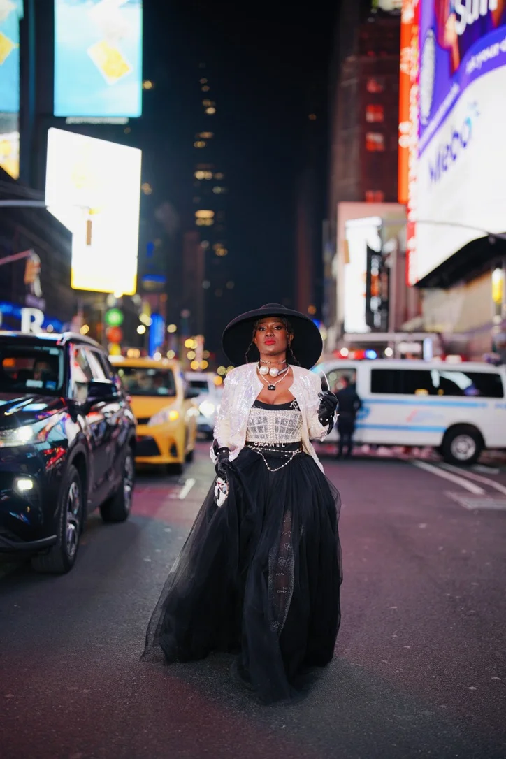 Dede Chancelor in Times Square at night, wide-brimmed hat and black tulle gown — the world outside the atelier.